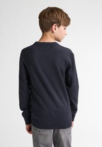 Pulover bleumarin cu mâneci lungi, guler și manșete canalate, având o textură netedă. Purtați cu pantaloni gri deschis, văzut din spate.