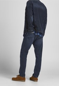 Chaqueta de mezclilla oscura con un aspecto desgastado, llevada sobre una camisa de cuadros azul, combinada con jeans de mezclilla oscura y zapatos de ante marrones. Conjunto casual.