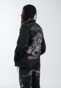 Chaqueta de satén negro con un gran gráfico de un dragón gris con acentos rojos en la parte posterior. El nombre de la marca "Ed Hardy" está bordado encima del diseño.