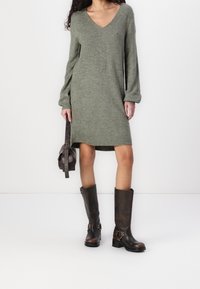 Abito lavorato a maglia verde oliva con scollo a V e maniche lunghe. Abbinato a stivali di pelle marroni al ginocchio con fibbie metalliche. Borsa marrone.