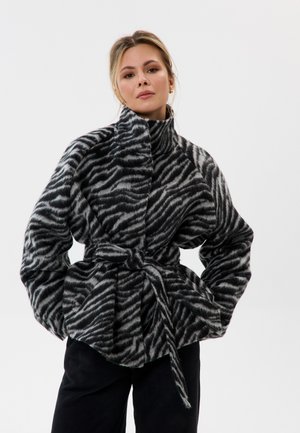 Frau trägt eine taillierte, hochgeschlossene Kunstpelzjacke mit Zebramuster, langen Ärmeln und schwarze Hosen vor einfachem Hintergrund.