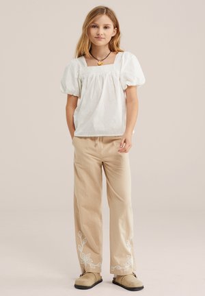 Mädchen mit langen blonden Haaren trägt ein weißes Oberteil mit Puffärmeln, beige bestickte Hose, beige Slipper und eine Blumenkette vor neutralem Hintergrund.