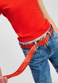 Sarkans jostas ar reljefu logo, valkāta kopā ar augsti jostasvietā sēdošām zilo džinsu biksēm. Gluda auduma un izteikta sprādzes aizdare. Ikdienas apģērba detaļa.