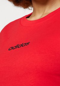 Crveni pamučni majica s okruglim izrezom i kratkim rukavima. Na prednjoj strani nalazi se crni "adidas" logo. Glatka tekstura.