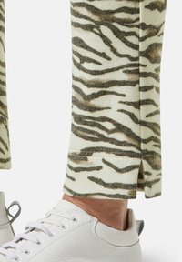 Tapered broek in crème met een zwart en bruin tijgerstrepenpatroon, met een split aan de onderkant, gecombineerd met een witte gestructureerde sneaker.
