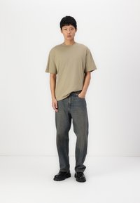 Beige cotton t-shirt with short sleeves, paired with loose blue denim jeans. Model wears black sturdy shoes, arms relaxed at sides.
