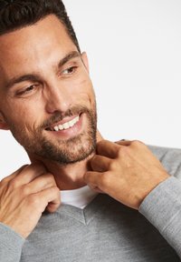Maglione grigio a v della morbida maglina, con una maglietta bianca sovrapposta visibile al colletto, caratterizzato da una vestibilità comoda e un design minimalista.