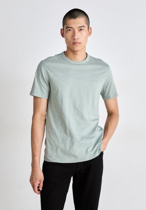 T-shirt vert clair à manches courtes en tissu doux. Doté d'un col rond classique et d'un logo subtil sur la poitrine. Associé à un pantalon noir.