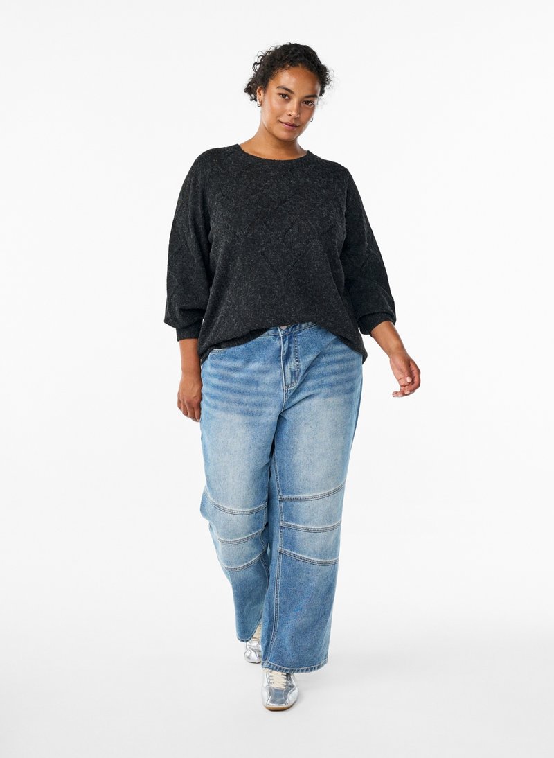 Kvinde går fremad iført en løs mørkegrå sweater, brede lyseblå jeans med knæsyninger og sølvfarvede sneakers mod en hvid baggrund.