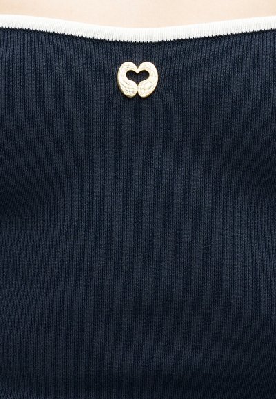 Χρυσό καρφίτσα σε σχήμα καρδιάς με δύο χέρια που φορούν μπλε μπλουζάκι με ραβδώσεις και λευκή επένδυση κατά μήκος του λαιμού.