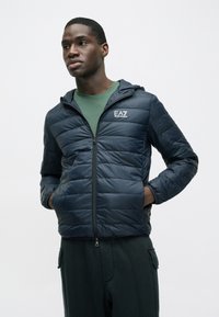 Tengerészkék steppelt dzseki cipzáras lezárással és kapucnival. Függőleges steppelt mintázattal és EA7 logóval a mellrészen. Zöld ing fölött viselve.