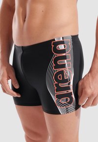 Schwarze Badeshorts mit weißen Wellenlinien und einem orangen "arena"-Logo an der Seite. Aus elastischem Material für Komfort und Leistung gefertigt.