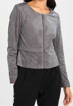 Femme portant une veste en daim grise ajustée à fermeture éclair avec manches longues et un pantalon noir, posant devant un fond uni.