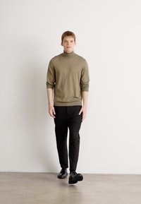 Olive zaļš turtlenecks džemperis, melnas slim-fit bikses un melni apavi, stāvot pretī gaišai fona ar gludu grīdu.