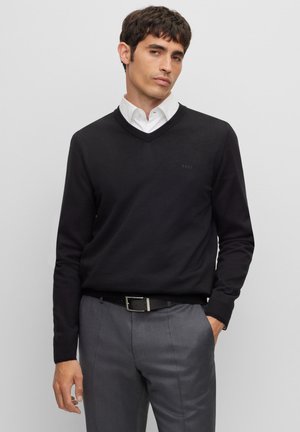 Zwarte v-hals trui gemaakt van gladde stof, met een subtiel logo op de borst. Gecombineerd met grijze broek en een zwarte riem.