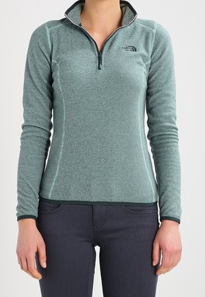 Kobieta ubrana w jasnoniemnozieloną polarową bluzę z suwakiem do ćwiartki na długi rękaw i ciemne dopasowane spodnie, stojąca z ramionami swobodnie opuszczonymi wzdłuż ciała.