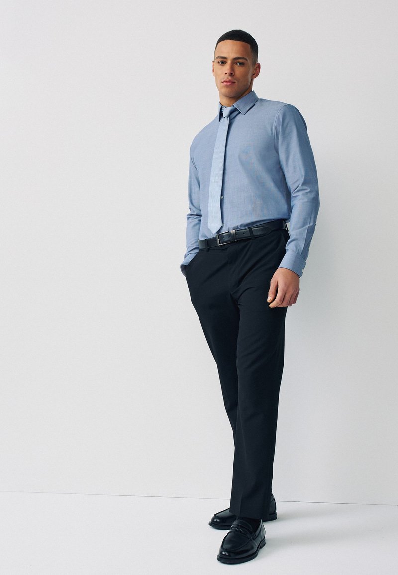 Homme debout portant une chemise habillée bleu clair, une cravate assortie, une ceinture noire, un pantalon noir et des chaussures habillées noires sur un fond clair uni.