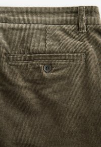 Gros plan sur la poche arrière d'un pantalon en velours côtelé marron avec une fente horizontale et un seul bouton foncé sous la passants de ceinture.