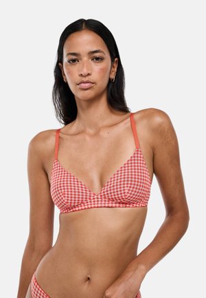 Femme aux longs cheveux foncés portant un haut de bikini à carreaux rouges et blancs sur un fond clair uni