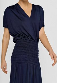 Navyblauwe jurk met een V-hals, korte mouwen en een geraffineerde taille. Soepele stof met een vloeiende onderkant, voorzien van geplooide textuur.