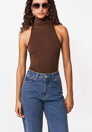 Kobieta o kręconych włosach, ubrana w brązowy, bezrękawnik z golfem, niebieskie, wysokowalne dżinsy i duże okulary przeciwsłoneczne, stojąca na białym tle.