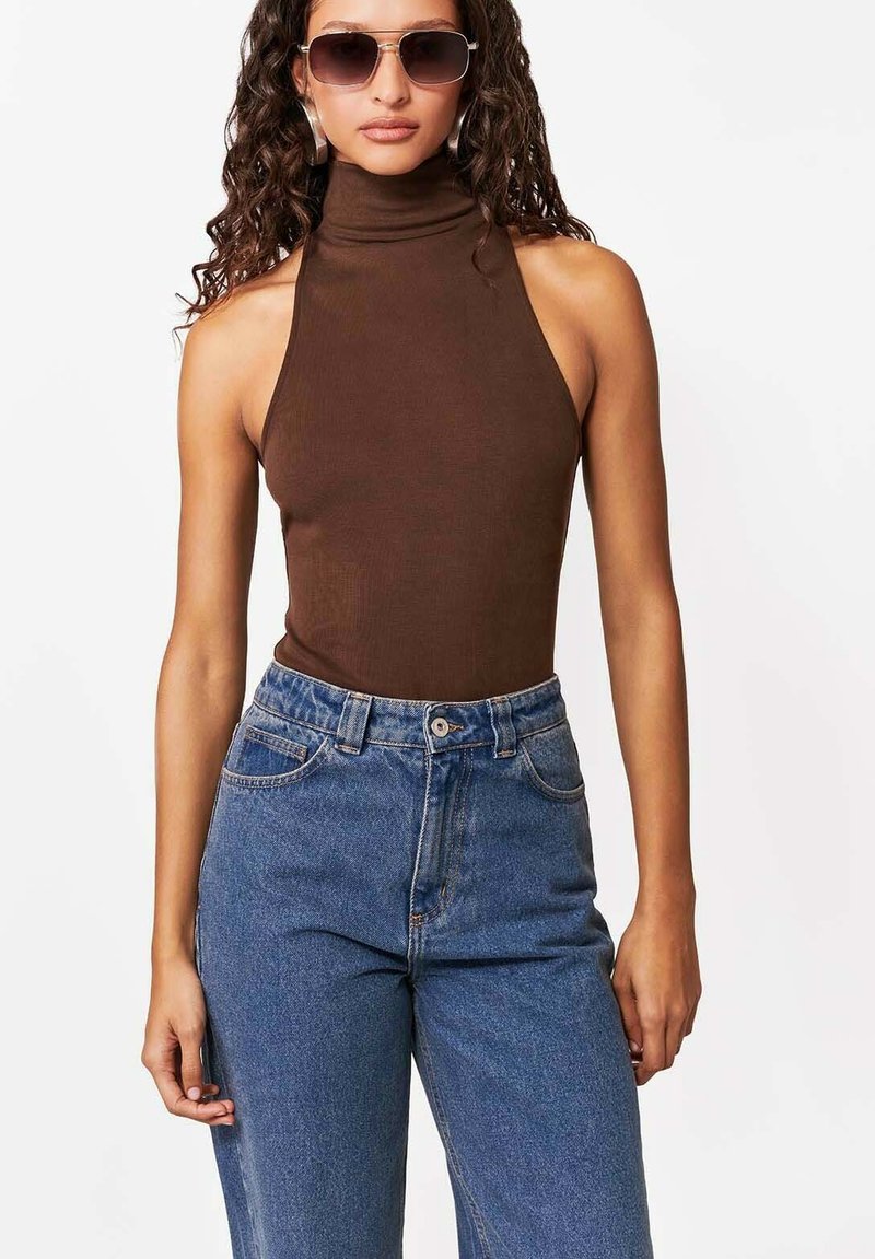 Kobieta o kręconych włosach, ubrana w brązowy, bezrękawnik z golfem, niebieskie, wysokowalne dżinsy i duże okulary przeciwsłoneczne, stojąca na białym tle.