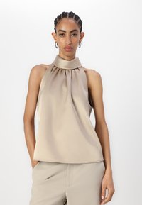 Top beige a collare alto in tessuto morbido con davanti plisettato. Abbinato a pantaloni beige chiaro. Design minimalista.