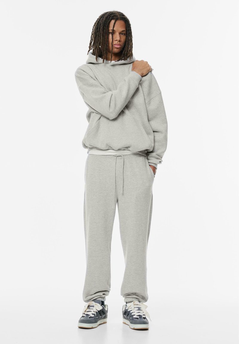 Grijze hoodie met een losse pasvorm, sweatpants met verstelbare tailleband en blauwe sneakers. Het materiaal lijkt zacht met een ontspannen, casual ontwerp.