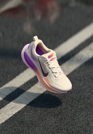 Vaaleanbeige ja violetti lenkkikenkä, jossa on tummanvärinen swoosh-logo, ilmassa asfalttitien päällä valkoisten kaistamerkintöjen yllä.