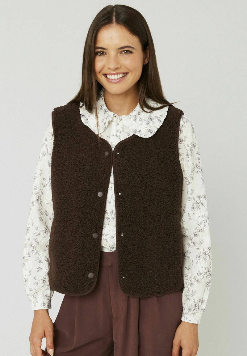 Gilet en polaire marron avec un col arrondi, des boutons-pression à l'avant et une texture douce, associé à une blouse fleurie et un pantalon foncé.