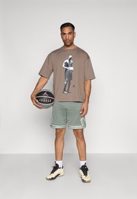 Pruun graafiline t-särk, millel on korvpallimängija print, rohelised lühikesed püksid valge äärisega, hoiab musta ja halli korvpalli, valged sokid ja kingad.