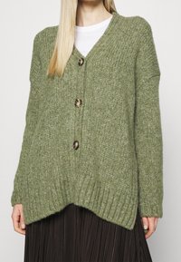 Green knitted cardigan featuring a V-neck, three large buttons, ribbed hem, and dropped shoulders, with a loose, relaxed fit.