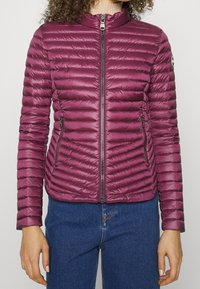 Figurvänlig, vinröd puffjacka med horisontellt quiltat mönster, hög krage, framzip och två zip-fickor på sidorna.