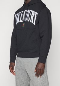 Sudadera negra con "NIKECOURT" en letras blancas en negrita, cuenta con un bolsillo frontal y capucha con cordón, combinada con pantalones deportivos grises.