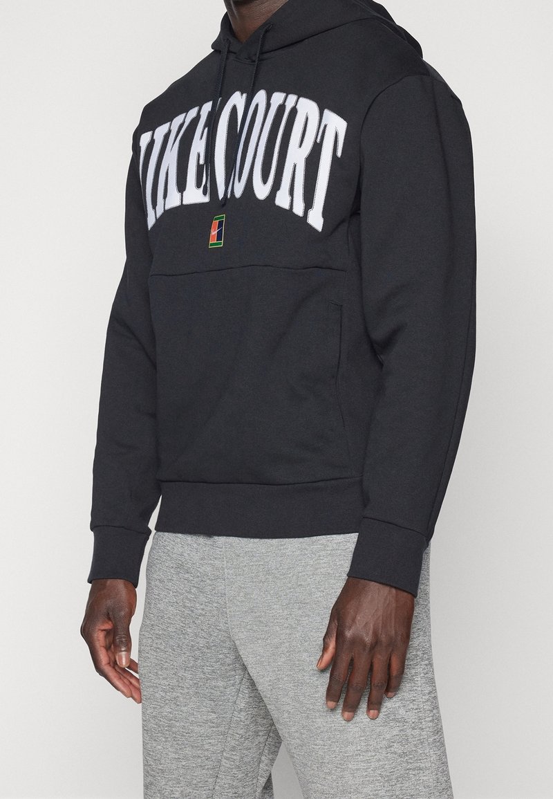 Sudadera negra con "NIKECOURT" en letras blancas en negrita, cuenta con un bolsillo frontal y capucha con cordón, combinada con pantalones deportivos grises.
