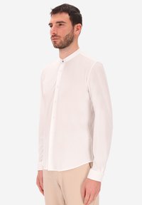 Uomo con capelli e barba scuri, che indossa una camicia bianca con colletto alla mandarina e pantaloni beige, in piedi contro uno sfondo chiaro e uniforme.