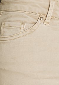 Beige denimtyg med subtil textur; har en framficka och en liten metallknappaccent nära midjan.