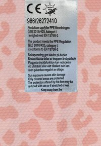 Etikett med säkerhets- och skötselråd på strukturerad tygbakgrund med ett rosa mönster. Inkluderar detaljer om överensstämmelse och UV-skydd.