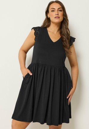 Frau trägt ein schwarzes ärmelloses Kleid mit gezackten Lochspitzen-Schultern und Taschen, posiert vor einem neutralen Hintergrund.