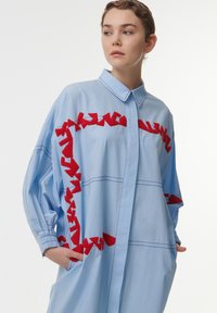 Lichtblauw oversized shirt met rode geborduurde accenten. Beschikt over een puntig kraag, knoopsluiting en zijzakken. Zacht stof en relaxte pasvorm.