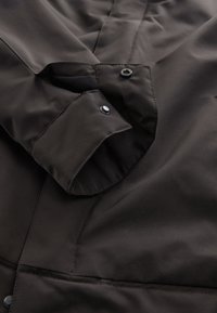 Közeli kép egy sötétbarna kabát mandzsettájáról, egyetlen patenttal záródik, sima szöveten jól látható varrási részletekkel.