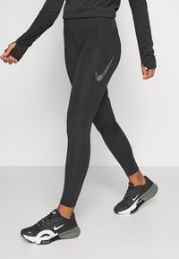 Černé atletické legíny s přiléhavým střihem, se reflexní Nike logem na boku, v kombinaci s černými a šedými běžeckými botami.