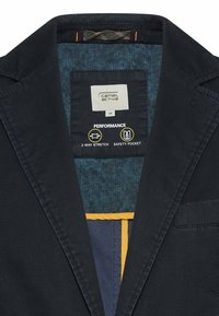 Nærbillede af navy blazer-krave med Camel Active størrelse 48 label med ydeevnefunktioner for 2-vejs stretchstof og sikkerhedslomme.