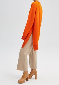 Pomarańczowy dziergany sweter o luźnym kroju z bocznymi rozcięciami, zestawiony z beżowymi spodniami o szerokich nogawkach oraz brązowymi botkami na masywnym obcasie.