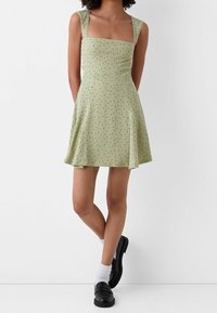 Vestito verde a coste con scollo quadrato, corpino aderente e gonna svasata. Macchie di punti neri su tutta la superficie. Indossato con scarpe nere e calzini bianchi.