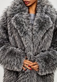 Fuskepelsjakke i grå nuancer med en tyk, fluffy struktur, brede revers og store pelsforede ærmer. Bemærkelsesværdig for sit luksuriøse udseende.