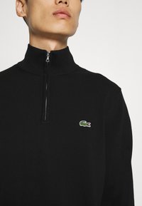 Pulover negru cu fermoar, cu textură ribbată, având un logo distinctiv verde de crocodil pe partea stângă a pieptului. Guler înalt și închidere cu fermoar rezistent.
