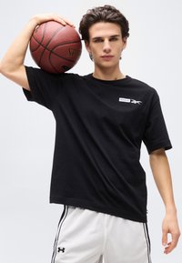 Νέος άνδρας με μαύρο μπλουζάκι Reebok και λευκά σορτς από δίχτυ, κρατώντας μια μπάλα του μπάσκετ Spalding στον ώμο του, μπροστά σε απλό φόντο.