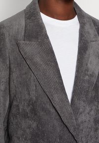 Blazer in velluto a coste grigio con righe verticali pronunciate, rever a punta e texture morbida, indossato sopra una T-shirt bianca.