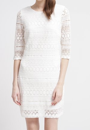 Mujer con un vestido blanco de crochet hasta la rodilla y mangas tres cuartos, sobre un fondo liso.